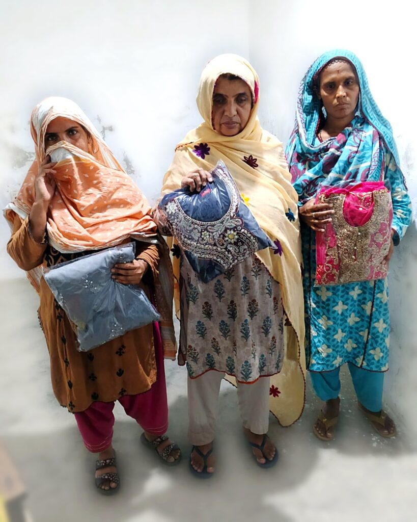 Three women in Larkana holding new embroidered suits donated through PHWO