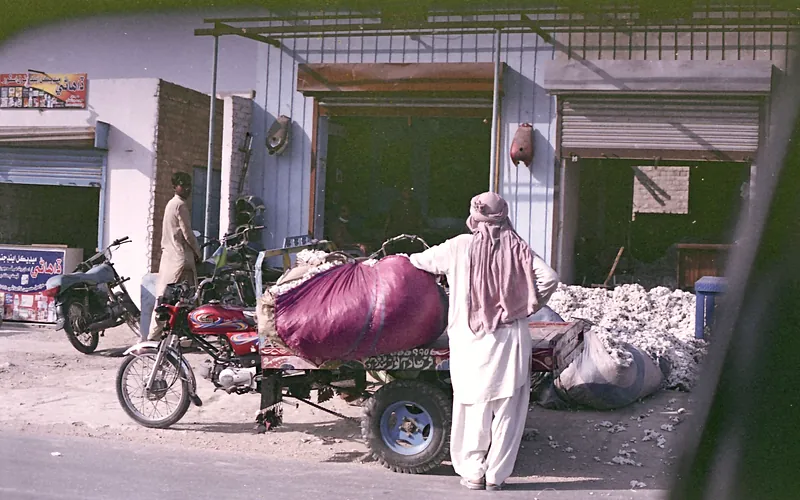 A man in traditional clothing stands by a motorcycle cart loaded with goods in Tando Ādam, Pakistan, Sindh