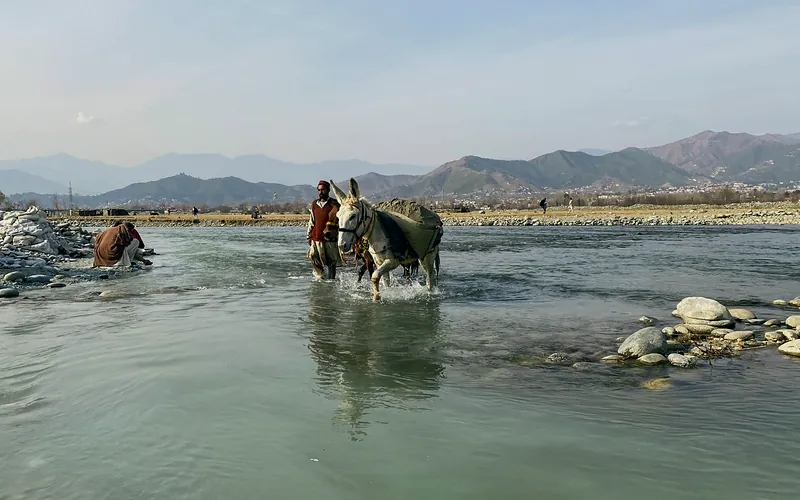 A man and donkey cross the scenic Swat River surrounded by mountains in Pakistan, Khyber Pakhtunkhwa