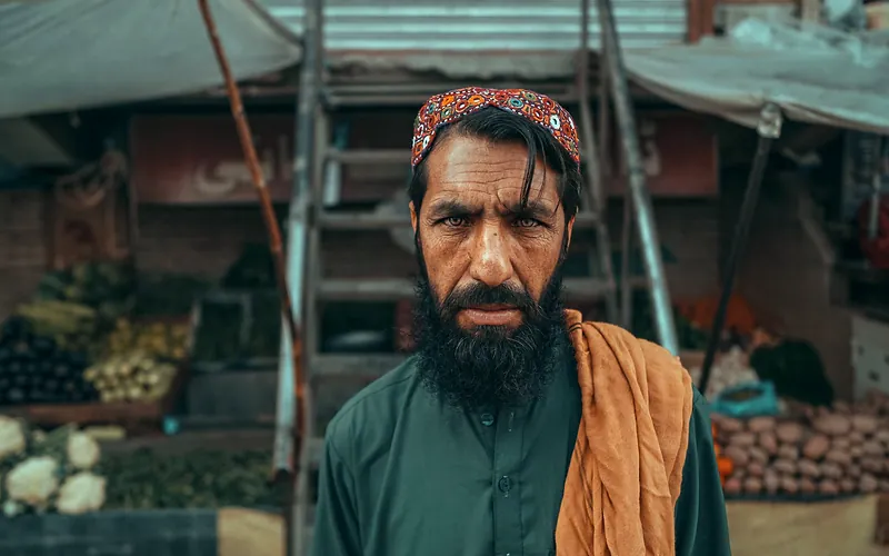 A bearded man in traditional attire stands in front of a market in Mansehra, Pakistan