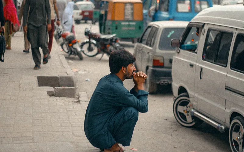 A man squats by roadside in urban Peshawar, Pakistan, displaying everyday life and bustling traffic, Khyber Pakhtunkhwa